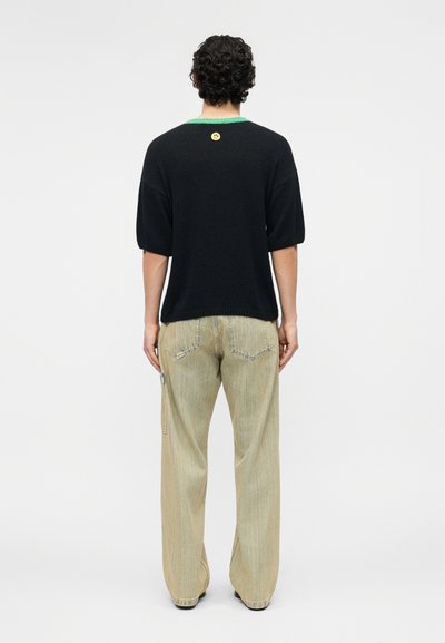 Άνδρας με σγουρά μαλλιά φοράει μαύρο πουλόβερ με κοντά μανίκια και πράσινο γιακά, μαζί με μπεζ παντελόνι φαρδύ, στέκεται με την πλάτη προς το φακό μπροστά σε λευκό φόντο.