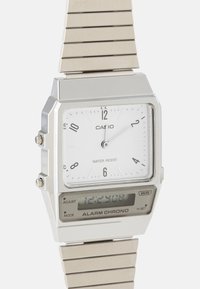 Montre Casio argentée avec un cadran analogique rectangulaire blanc comportant des chiffres arabes, une indication de résistance à l'eau et un affichage numérique sur un bracelet métallique.