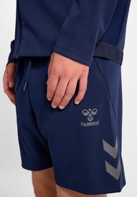 Marineblaue Sportshorts aus glattem Stoff, mit einem Logo und geometrischen Akzenten in einem helleren Grau, ausgestattet mit einem elastischen Bund und Kordelzügen.