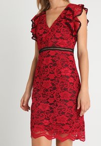 Robe en dentelle rouge avec un col en V, des manches à volants et une ceinture noire. Motif floral, ourlet festonné et texture transparente sur l'ensemble.