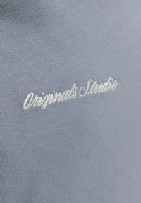 Jasnoniebieska tkanina z białym, haftowanym napisem "Originals Studio" kursywą, ze szczególnym uwzględnieniem faktury i detali szwu.