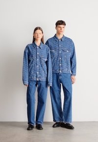 Dvije plave traper jakne i hlače širokih nogavica s teksturiranim uzorkom. Oba odjevna predmeta imaju dvostruke džepove na prsima i zatvaranje na kopče.