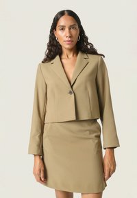 Klippt beige kavaj med enstaka knappstängning, i kombination med en matchande figursydd kjol, slät textur och långa ärmar.