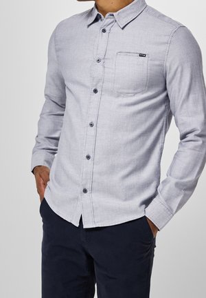 Man draagt een lichtgrijs overhemd met knopen en een borstzak en donkerblauwe broek, handen in de zakken, tegen een effen achtergrond.