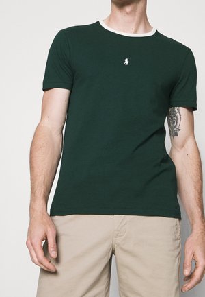 Mężczyzna w ciemnozielonej koszulce z krótkim rękawem z białym wykończeniem i małym białym logo polo, beżowe szorty oraz kwiatowy tatuaż na lewym ramieniu.