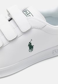 Weiße synthetische Sneakers mit Klettverschluss, grünem gesticktem Pferdelogo und strukturiertem Sohlenprofil mit dezenten Rillen.
