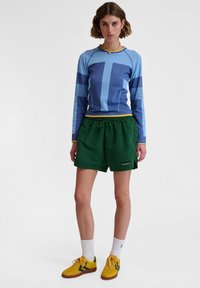 Långärmad blå tröja med grafisk design, kombinerad med gröna shorts med elastisk midja och logotyp. Gula sneakers och vita strumpor fulländar outfiten.