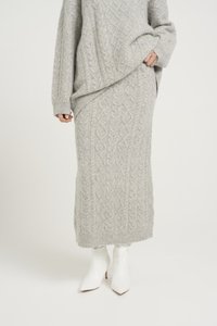 Un pull en laine gris clair tricoté et une jupe assortie avec un motif en maille torsadée. Le pull a une coupe oversize ; la jupe est longue jusqu'à la cheville. Bottes à talons blanches.