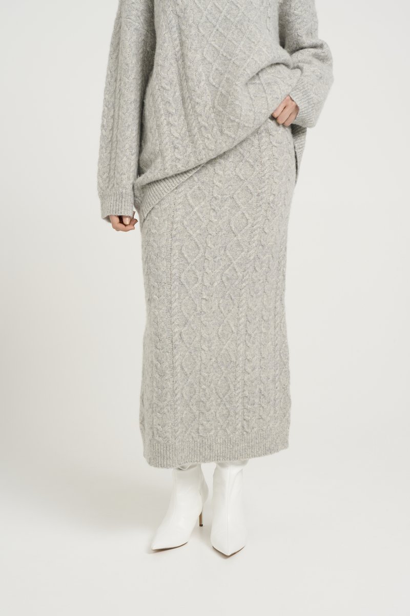 Un pull en laine gris clair tricoté et une jupe assortie avec un motif en maille torsadée. Le pull a une coupe oversize ; la jupe est longue jusqu'à la cheville. Bottes à talons blanches.