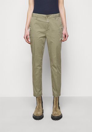 Pantalones de corte ajustado color caqui combinados con botines de ante marrón claro y una blusa negra metida por dentro, vistos desde la cintura hacia abajo.