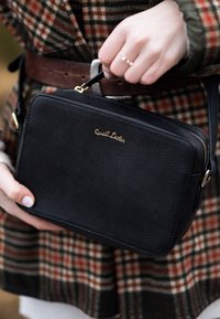 Crna teksturirana crossbody torba s zlatnim logom, zatvaranjem na patentni zatvarač i podesivim remenom, koju drži osoba u outfitu s kariranim uzorkom.