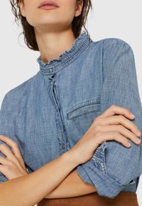 Camicia in denim blu chiaro con collo a volant, chiusura a bottoni e una piccola tasca sul petto. Le maniche sono arrotolate, evidenziando la texture.
