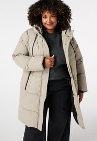 Manteau matelassé beige avec capuche, poches latérales et éléments en métal noir. Design épais, matelassé, avec un ourlet plus long et une coupe décontractée.