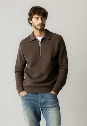 Mann trägt braunen langärmeligen Reißverschluss-Pullover und hellblaue Jeans, steht mit Händen in den Taschen.