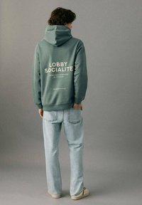 Sudadera verde con capucha y un bolsillo tipo canguro, con texto blanco en la espalda. Combinada con vaqueros azul claro y zapatillas casuales.