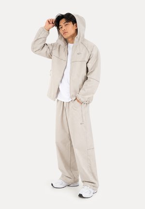 Jeune homme portant une veste à capuche beige clair et un pantalon large assorti avec des baskets blanches, debout avec une main dans la poche.