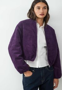 Lila Cordjacke aus Cord mit gerippter Textur, cropped Design und elastischen Bündchen. Reißverschluss vorne, kombiniert mit einem weißen Hemd.