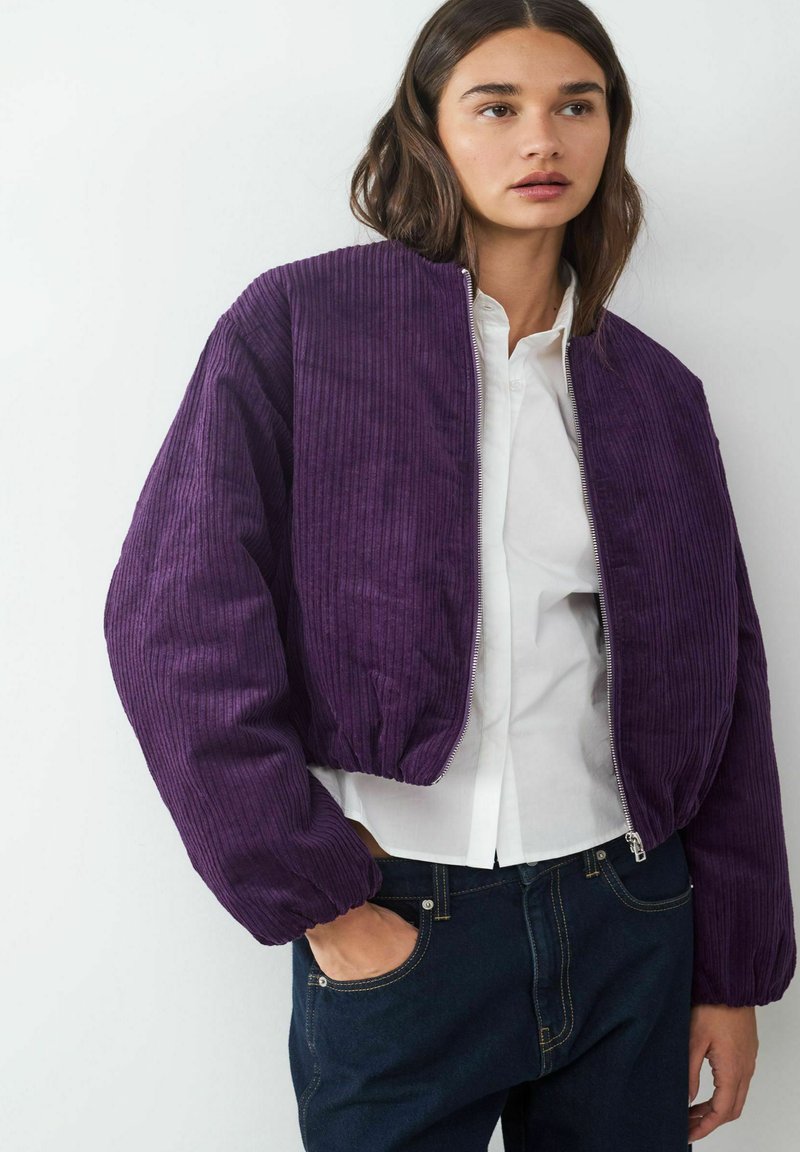 Lila Cordjacke aus Cord mit gerippter Textur, cropped Design und elastischen Bündchen. Reißverschluss vorne, kombiniert mit einem weißen Hemd.