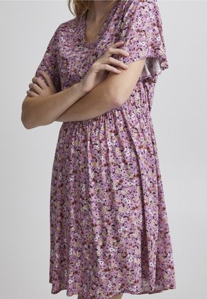 Osoba w liliowej sukience w kwiaty z krótkimi rękawami, z założonymi ramionami, stojąca na tle jasnoszarym jednolitym.