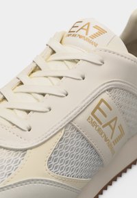 Λευκό αθλητικό παπούτσι Emporio Armani EA με πάνελ από δίχτυ, εκρού λεπτομέρειες και χρυσό λογότυπο στη γλώσσα και στο πλάι.