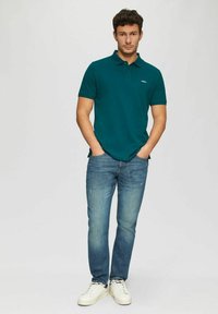 Türkises Polohemd mit klassischem Kragen, kombiniert mit hellblauen Jeans. Weiße Sneakers vervollständigen das Outfit. Einfaches Design, keine Muster.