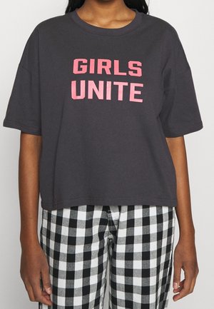 Sort oversized T-shirt med pink "GIRLS UNITE" tekst båret med sort og hvid ternede bukser på en person med medium hudtone.