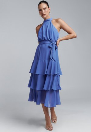 Blaues Chiffonkleid mit Halterhals, taillierter Oberteil und drei Lagen Rüschen am Rock, mit einem gebundenen Taillengürtel. Absatzsandalen.