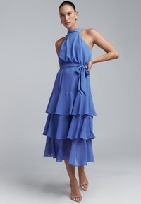 Plava čipkasta haljina s halter izrezom, prilagođenim korzetom i tri sloja nabranica na suknji, suženi struk s mašnom. Sandale na petu.