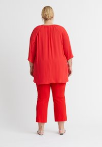 Rotes Oberteil mit fließenden Ärmeln und gerafftem Rücken, kombiniert mit roten cropped Hosen. Beide Kleidungsstücke haben eine glatte Textur und ein einfaches Design.