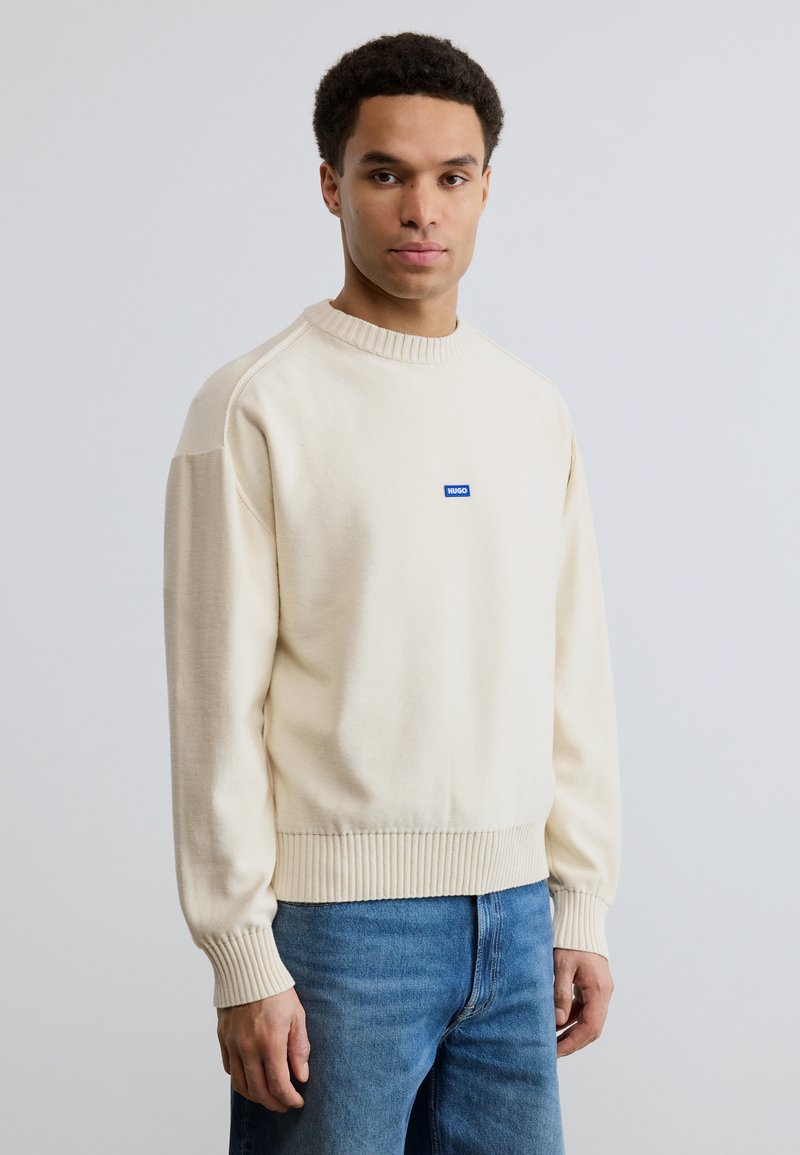 Homme portant un pull en tricot crème avec col et poignets côtelés, patch logo bleu et jean bleu, debout devant un fond clair uni.