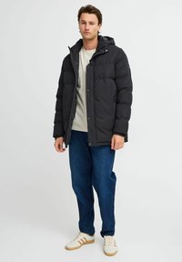 Svart puffjakke med hette, som har glidelås og knappelukking, to sidelommer og en quiltet tekstur, brukt over en lyse skjorte.