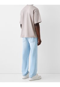 Man som bär en ljusbeige kortärmad skjorta, ljusblå löst sittande jeans och vita sneakers står mot en enfärgad vit bakgrund.