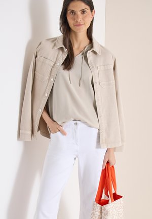 Femme aux longs cheveux noirs portant une veste beige sur un sweat à capuche clair et un pantalon blanc, tenant un sac fourre-tout beige à motif orange.
