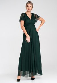 Langes, dunkelgrünes Kleid mit tailliertem Oberteil, transparenten kurzen Ärmeln und einem fließenden Tüllrock. Der Stoff ist glatt mit einem ausgewogenen Fall. Schwarze High Heels.