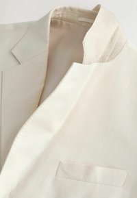 Blazer de couleur crème avec un revers cranté, une poche poitrine passepoilée simple et une doublure intérieure visible autour du col.