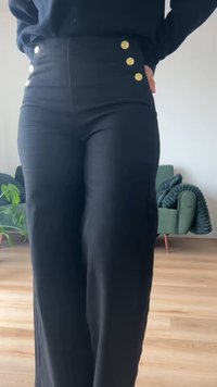 Γυναίκα που φοράει μαύρο παντελόνι ψηλόμεσο με τρία χρυσά κουμπιά σε κάθε πλευρά, στέκεται μέσα σε σπίτι με έναν πράσινο καναπέ και φυτό στο παρασκήνιο.