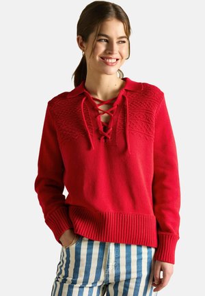 Red knit sweater featuring a lace-up neckline, textured design across the chest, and ribbed cuffs with a loose fit. Paired with striped pants.