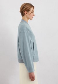 Light blue bomber jacket made from soft fabric, featuring a ribbed neckline, front pocket, and relaxed fit. Smooth texture.