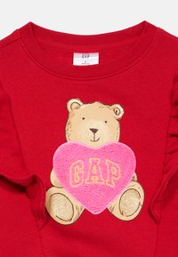 Czerwona bluza z nadrukiem niedźwiedzia z różowym, puszystym sercem z napisem "GAP". Falbaniaste rękawy dodają projektowi faktury.