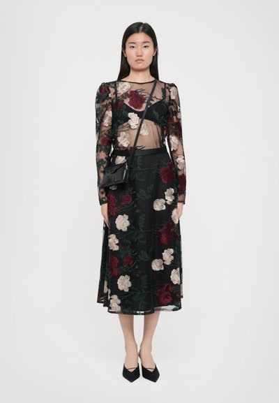 Rochie neagră transparentă cu broderie florală în roșu și crem, cu mâneci ajustate și fustă lungă. Asortată cu o poșetă mică neagră și tocuri.