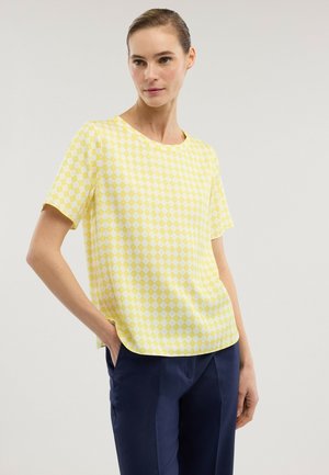 Frau trägt ein gelb-weiß kariertes kurzärmeliges Oberteil und dunkelblaue Hose, posiert mit einer Hand in der Tasche vor einem schlichten Hintergrund.