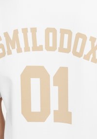 Witte katoenen t-shirt met de tekst "SMILODOX 01" in een taupe, vetgedrukt lettertype op de borst. Het ontwerp is eenvoudig en minimalistisch.