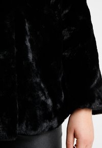 Schwarze Kunstpelzjacke mit einer weichen, plüschigen Textur, die einen lässigen Schnitt und weite Ärmel bietet. Der Stoff hat einen dezenten Glanz.