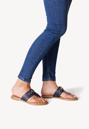 Ben i blå skinny jeans med sorte læder flade sandaler med gulddetalje, der står på en hvid overflade.