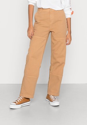 Person trägt beige weite Hosen und passende High-Top-Sneaker, steht vor einem schlichten hellen Hintergrund.