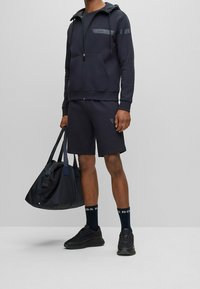 Marinblå zip-up hoodie med logotyp, matchande shorts, svarta sneakers och gymväska. Texturerad tyg, atletisk passform och subtila designdetaljer.