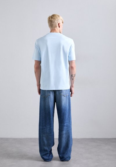Blond person stands facing a light gray wall wearing a light blue short-sleeve shirt and loose-fitting blue jeans on a gray floor.