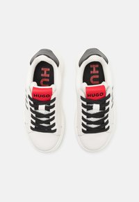 Hvite sneakers med svarte detaljer, med en rød etikett med "HUGO" på tungen, svarte snøringer og en teksturert innersåle.