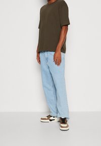 Person wearing olive green oversized T-shirt, light blue loose jeans, and beige and black sneakers standing against a plain white background.