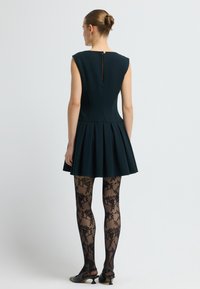 Robe sans manches de couleur marine avec une jupe plissée, un col rond et une fermeture éclair à l'arrière. Le modèle porte des collants à motifs noirs et des chaussures à talons noires.